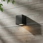 Aplique de pared MONACO moderno negro de hierro con luz cálida en pared de madera.