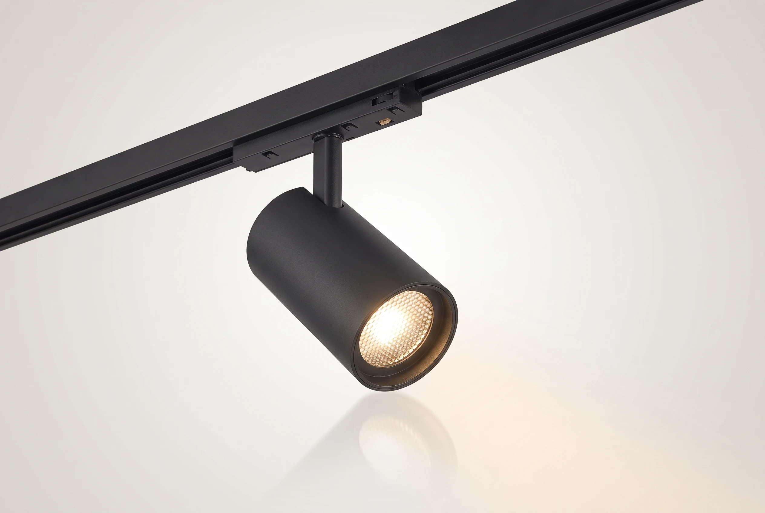 Spot orientable Smart de 26W montado en un tramo de riel magnético negro sobre fondo claro, cabezal cilíndrico inclinado con la lente encendida en luz cálida