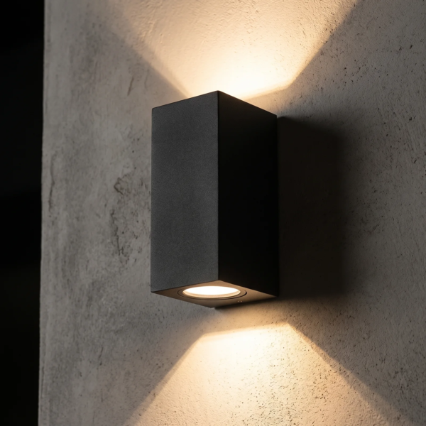 Detalle del aplique de exterior Vasastan negro mate mostrando el doble haz de luz cálida hacia arriba y hacia abajo sobre una pared de microcemento